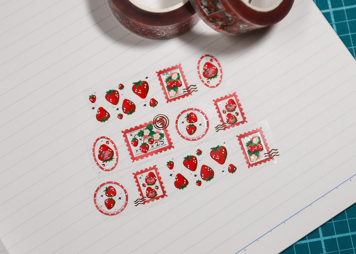 Strawberry-themed washi tape rolls and individual strips laid out on lined paper, with the rolls partially visible at the top and a green cutting mat edge visible on the right.