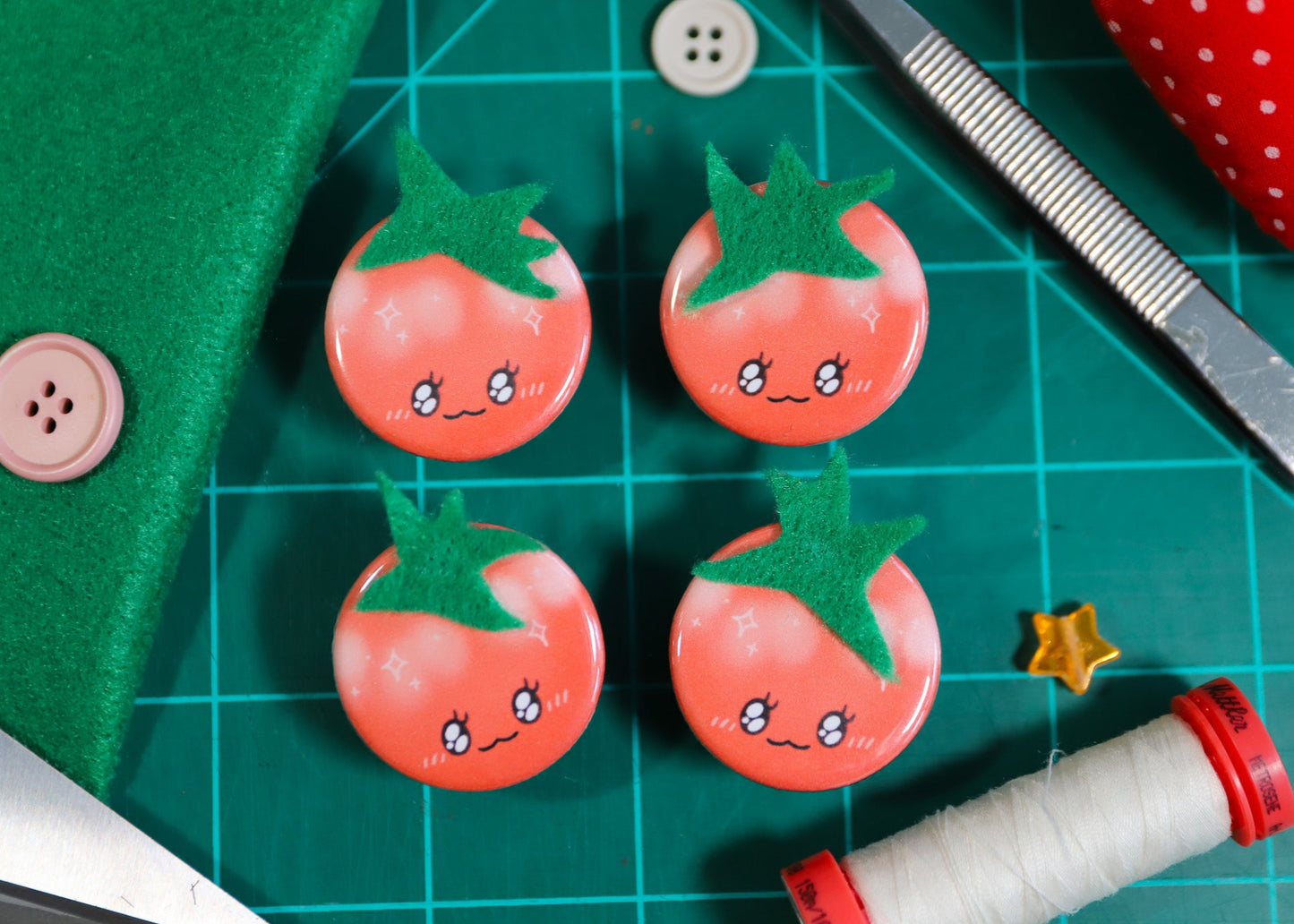 Four cute tomato-shaped buttons with green stems on a green cutting mat with sewing tools. Handmade tomato pins and magnets.
