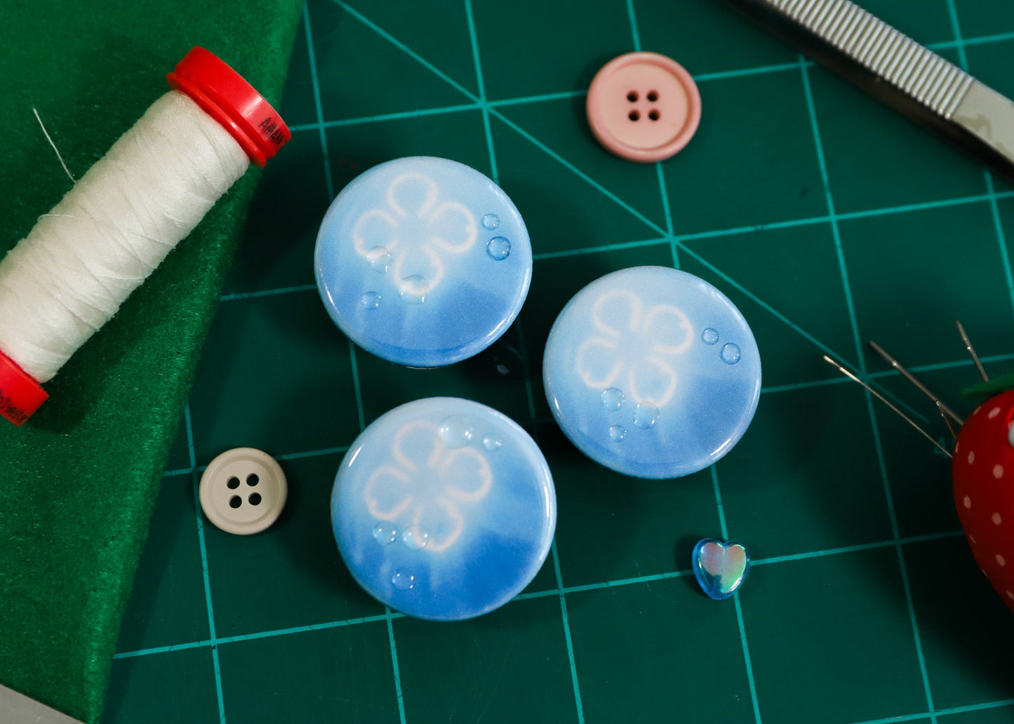 Blue buttons with white patterns on a green cutting mat with sewing tools. Moon Jelly fish pins and magnet with water drops on them.