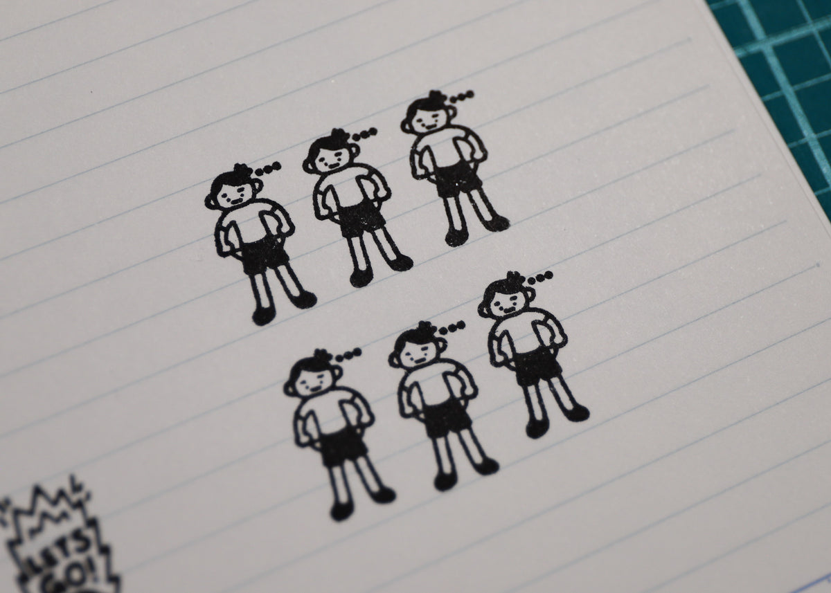A close-up of a lined notebook page with six black cartoon stick-figure drawings arranged in two rows. A green cutting mat is partially visible in the top-right corner.