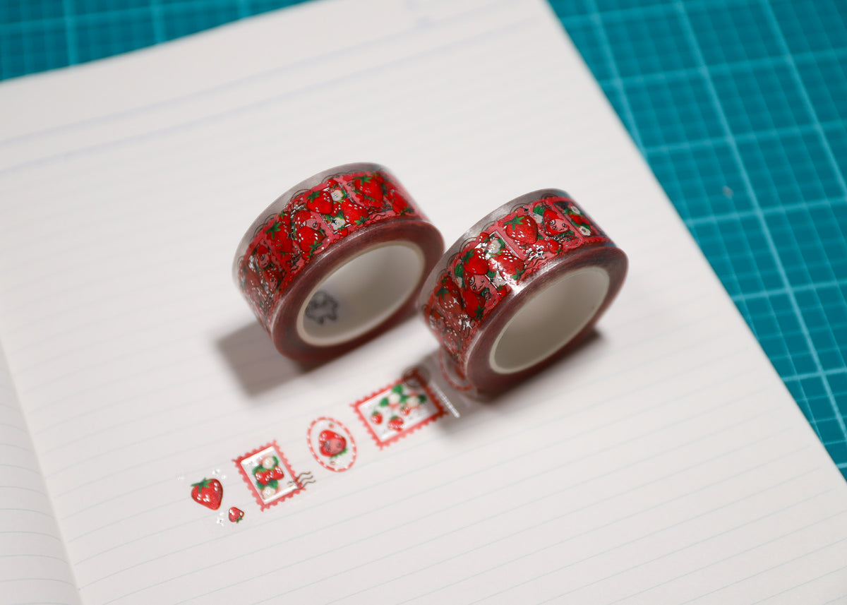 Two rolls of washi tape with a red and green strawberry pattern, displayed on a lined sheet of paper over a green cutting mat.