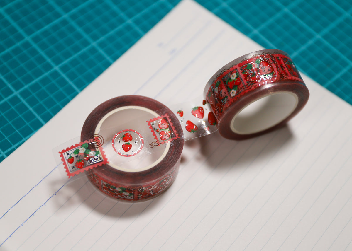 Two rolls of decorative washi tape with a red and green strawberry pattern, shown on lined paper with a teal cutting mat in the background.