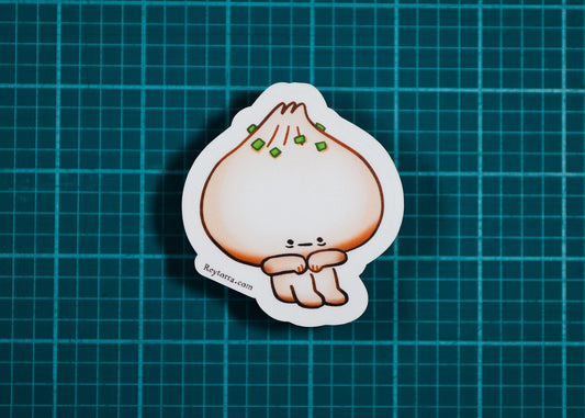 A die-cut sticker featuring a cute cartoon dumpling character with green garnish, placed on a dark teal grid cutting mat.
