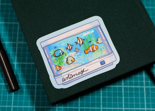 A dark green hardcover notebook with a rectangular sticker depicting colorful clownfish in an aquarium, placed on a green cutting mat next to a black pen.
