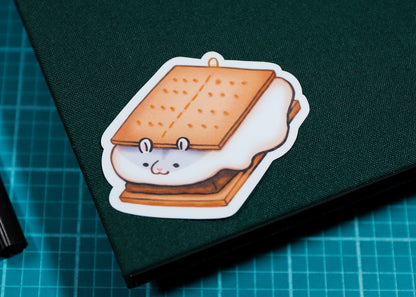 A cute cartoon s'mores die-cut sticker with a smiling face is shown on the dark green cover of a notebook, placed on a blue cutting mat next to a black pen.