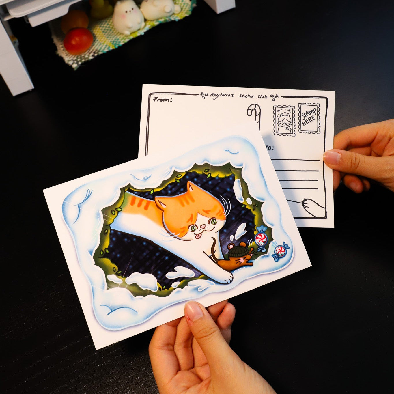 Two hands holding postcards with illustrations of cats on a dark surface.