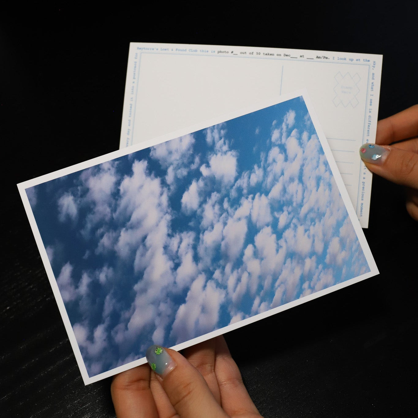 Hand holding a postcard with a blue sky and clouds design on a dark surface