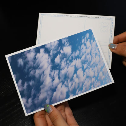 Hand holding a postcard with a blue sky and clouds design on a dark surface
