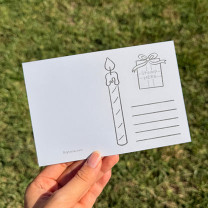 A hand holds a white postcard featuring simple black line drawings of a candle and a gift box, photographed outdoors against a blurred green grass background.