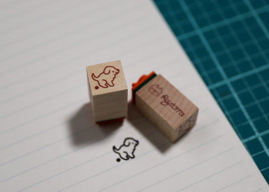 Two wooden rubber stamps with a dog design, one showing the stamp face and the other showing the branded end with orange ink pad, placed on lined paper next to a green cutting mat.