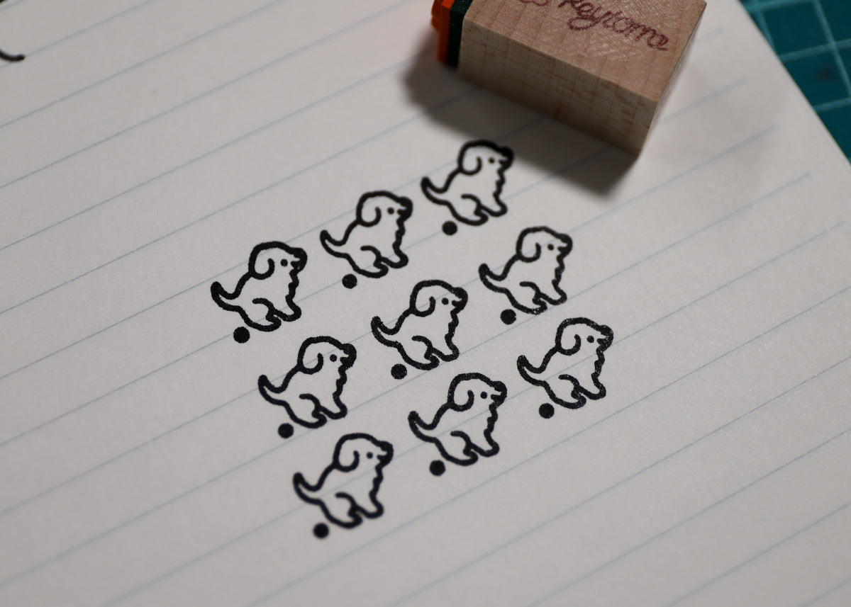 A wooden rubber stamp with a dog outline design is shown on lined paper, with several stamped dog outlines visible and part of the stamp block in the upper right corner.