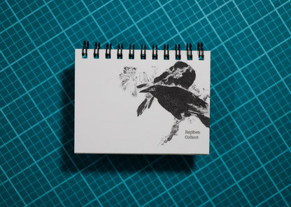 A spiral-bound notebook with a black-and-white bird illustration on the cover, placed on a teal grid cutting mat background.