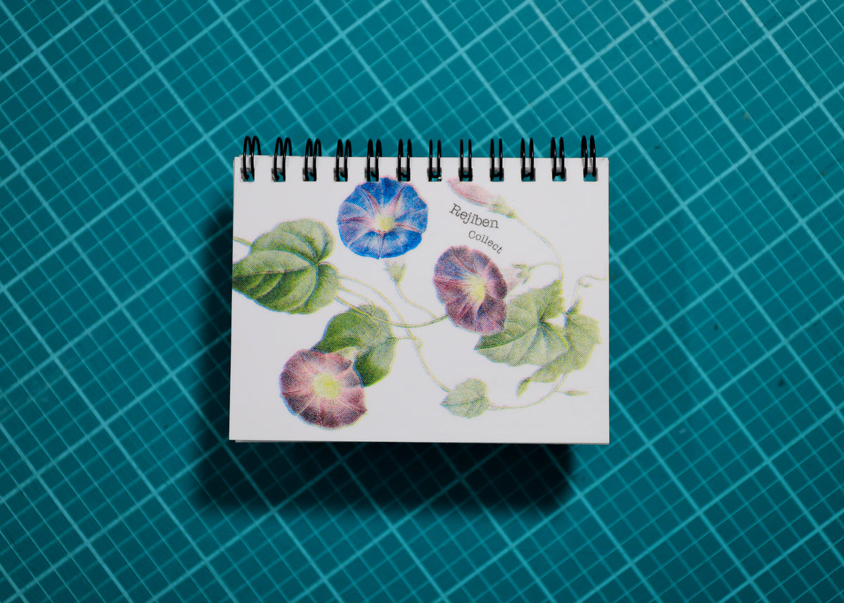 A small spiral-bound notebook or planner cover with a botanical floral print of blue and purple morning glories on a white background, placed on a teal cutting mat with a grid pattern.