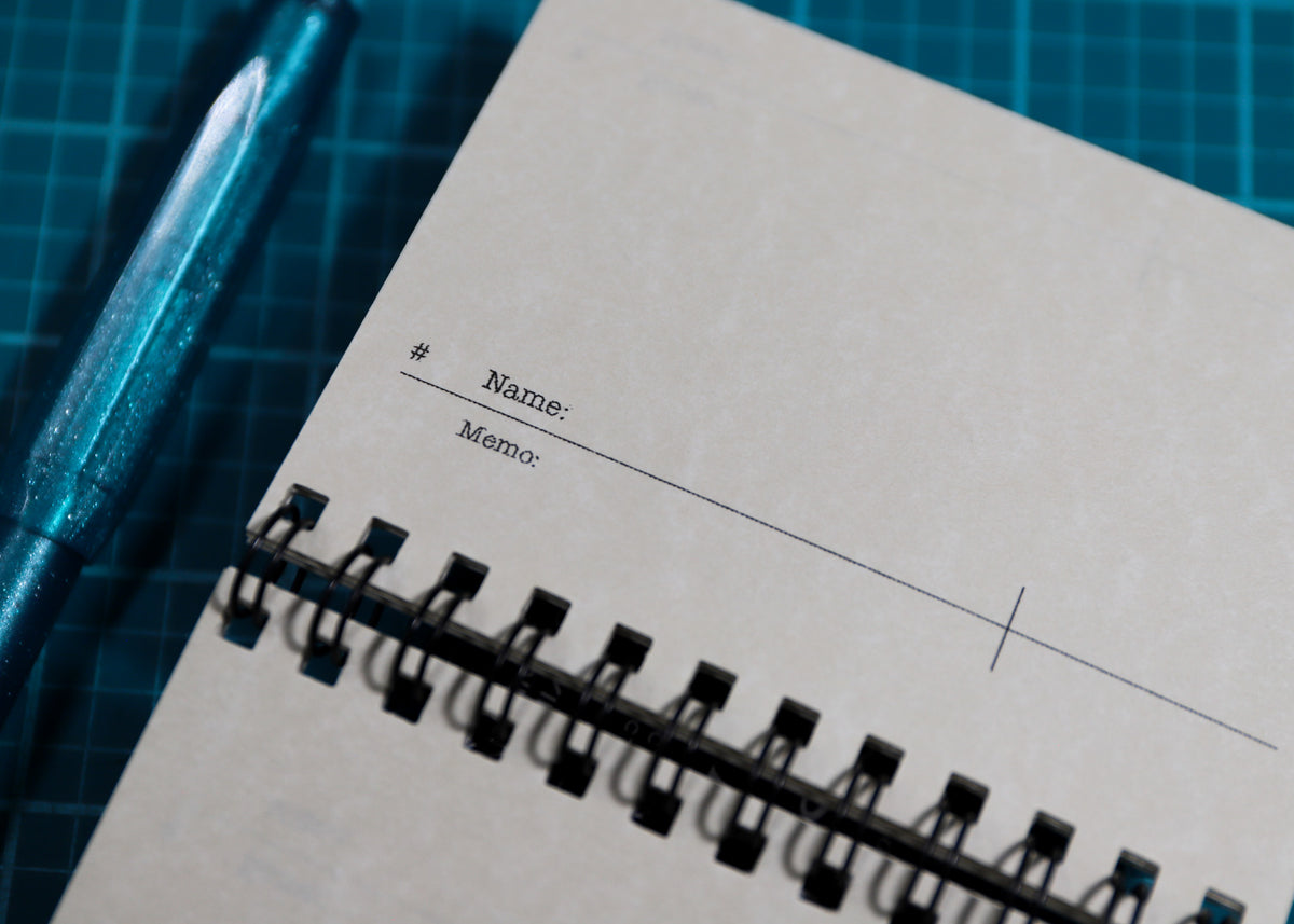 A close-up of a spiral-bound memo pad with blank lines for 'Name:' and 'Memo:' on a blue gridded cutting mat, with a metallic teal pen partially visible on the left.