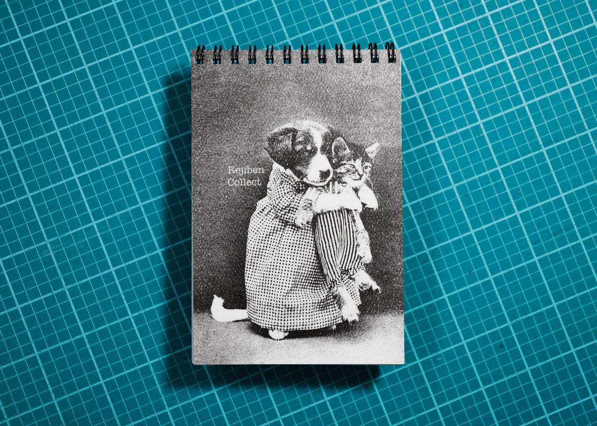 A spiral-bound notebook featuring a vintage black-and-white photograph of a dog and cat in clothing, placed on a teal cutting mat with a grid pattern.
