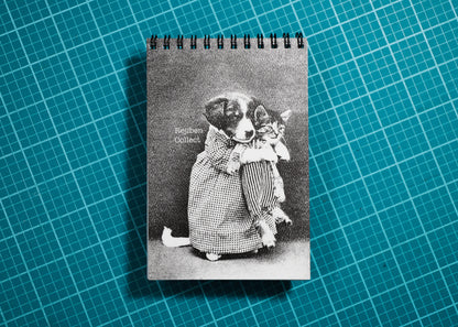 A spiral-bound notebook featuring a vintage black-and-white photograph of a dog and cat in clothing, placed on a teal cutting mat with a grid pattern.