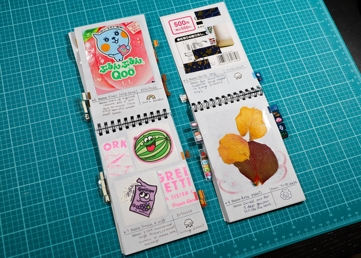 Two spiral-bound notebooks with Japanese illustrations and text, displayed on a green cutting mat background.